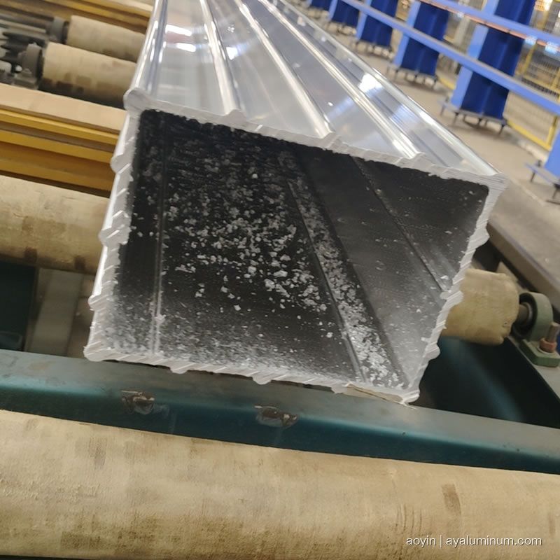 Cut end of a freshly extruded aluminum square tube on the runout rollers, showing the hollow section walls, internal ribs, and corner radii