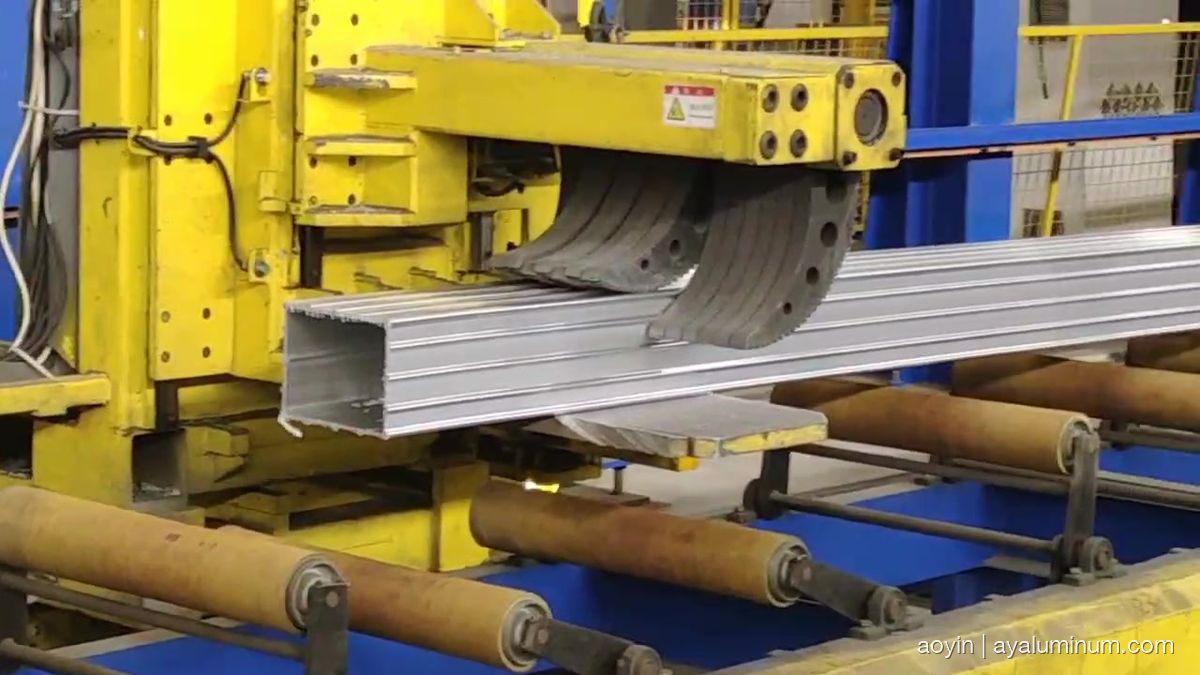 Hollow aluminum box profile emerging from the extrusion die under the press shoe onto the runout rollers, still hot